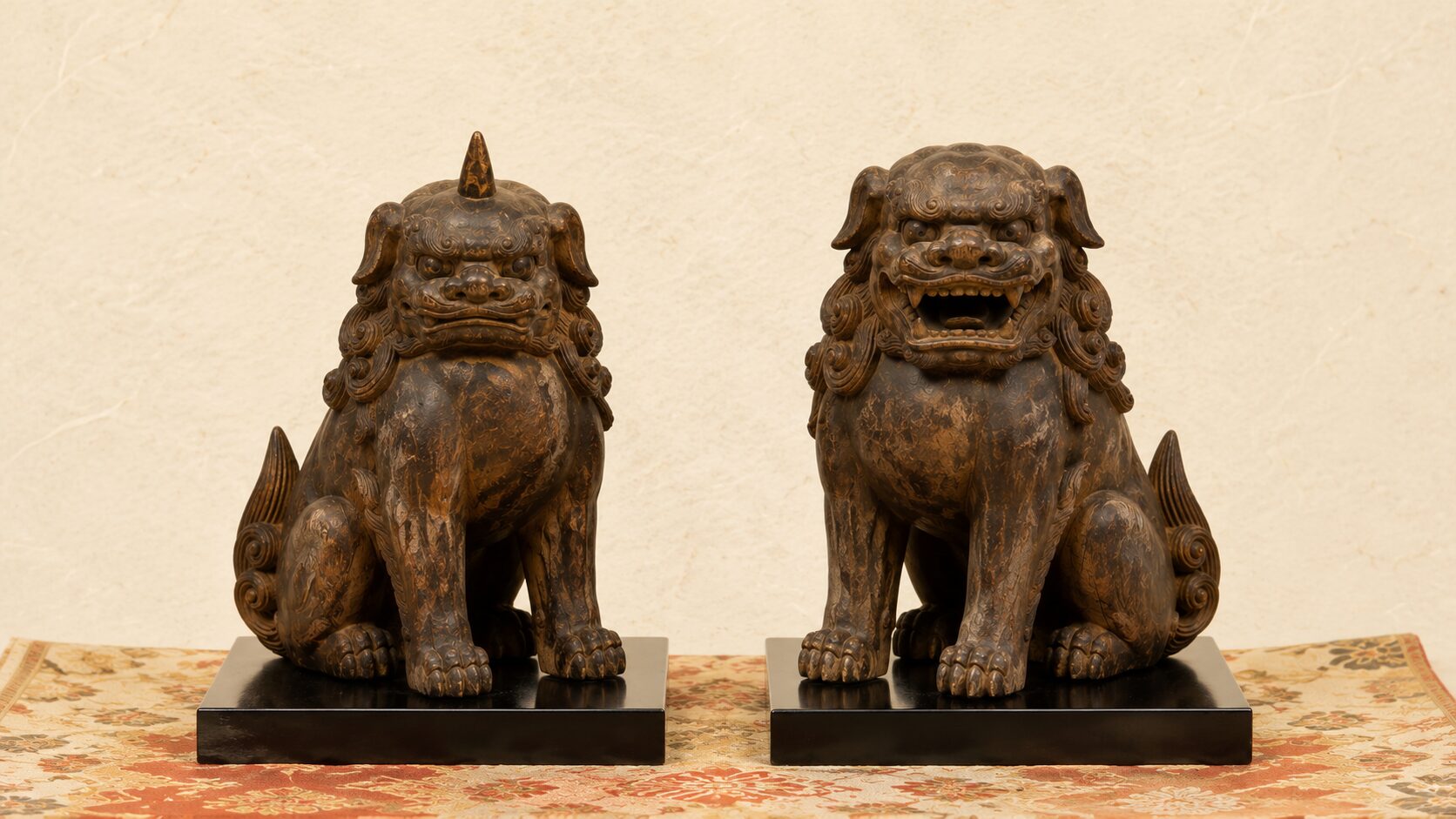 ivory washi 背景に painterly な木造獅子・狛犬一対の正面接写。中央に平安期の小型木造像(右が獅子=阿形・角なし、左が狛犬=吽形・頭に一本の角)、暗い飴色の古木、漆台座、几帳の布。平安時代の獅子・狛犬ペア構造を象徴