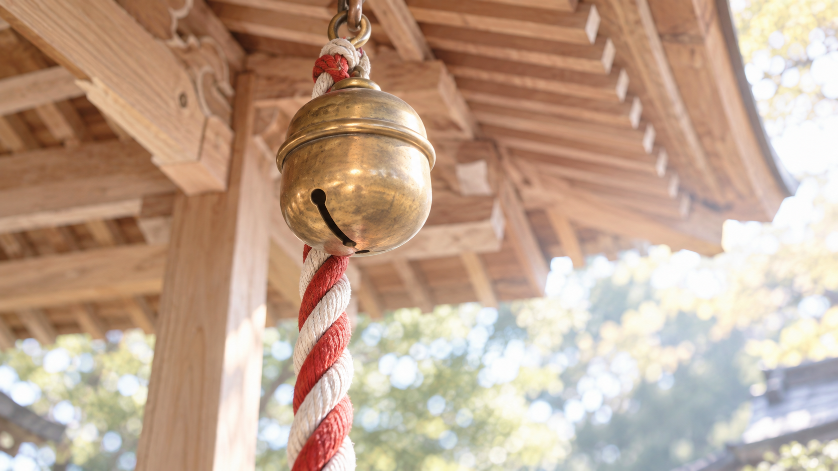 神社の拝殿の軒下から下がる丸い真鍮の鈴が1つと、朱と白の木綿を綯った鈴緒が1本を、少し下から見上げた接写画像。背景には白木の柱と軒の木組みがやわらかくぼけ、朝の光が差し込む。人の姿はない。神社でお参りやご祈祷を受ける場の象徴画像。